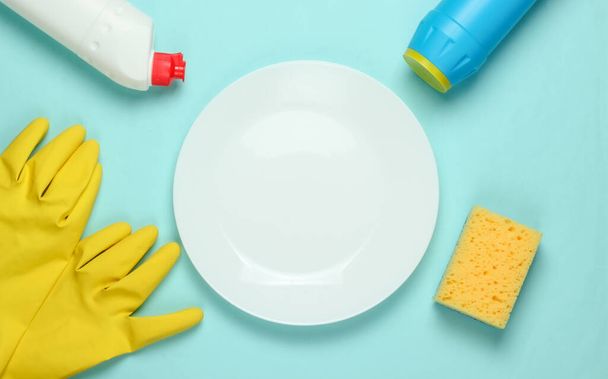 Washing dishes concept. Plate with a sponge and rubber gloves, bottle of detergent on a blue pastel background. Top view - Photo, Image