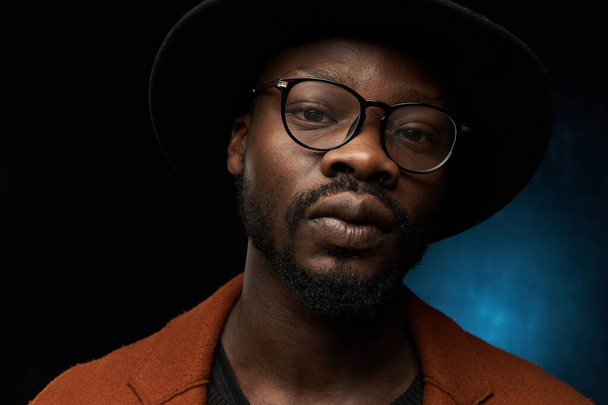 portrait photo of a black man with glasses, black hat, brown jacket and jacket on a dark background in the studio, who is looking thoughtfully at the camera - Фото, изображение