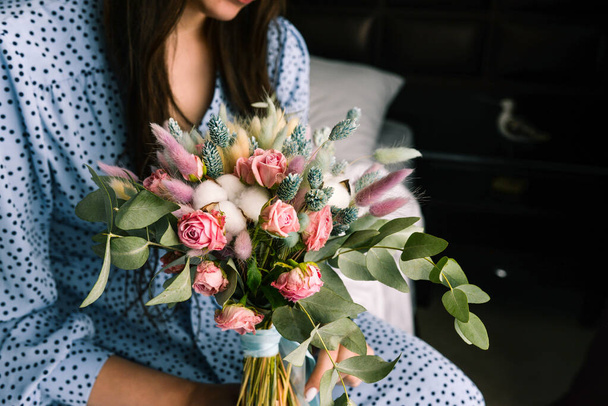 Girl with a bouquet of flowers close-up. Woman is sitting on a bed in a blue dress and holding dried flowers. Concept of wedding, relationships and love. - Photo, Image