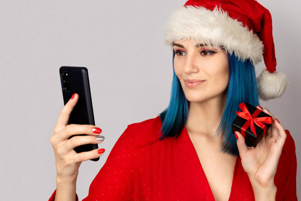 Gelukkige jonge vrouw in santa hoed met geschenkdoos en smartphone over grijze achtergrond. Kerstmis online shopping sale concept - Foto, afbeelding