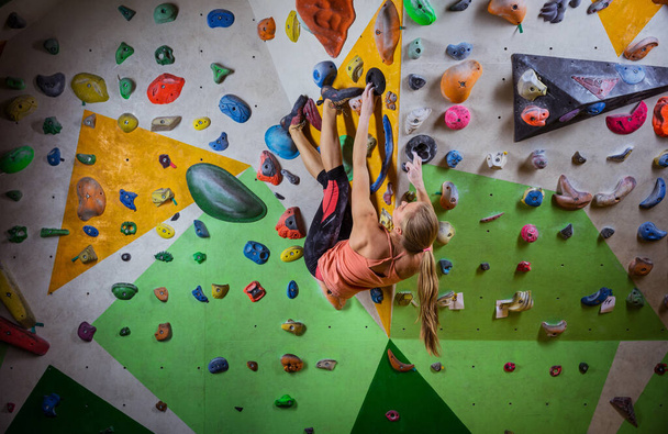 Молодая женщина боулдеринг на нависающей стене в крытом альпинистском зале, делая сложный шаг, чтобы достичь финального захвата - Фото, изображение