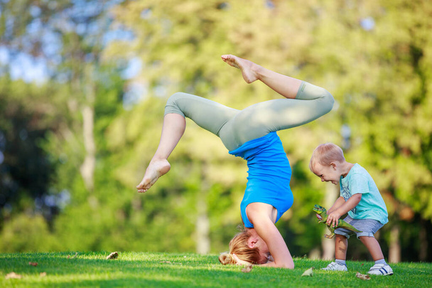 Young woman working out outdoors, standing on head, little son playing beside her - Fotografie, Obrázek