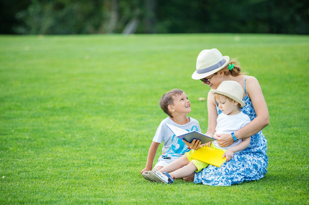 Joven madre leyendo libro a sus dos hijos pequeños mientras está sentada en el césped en el parque. Niños y madre disfrutando de un hermoso día de verano. - Foto, Imagen