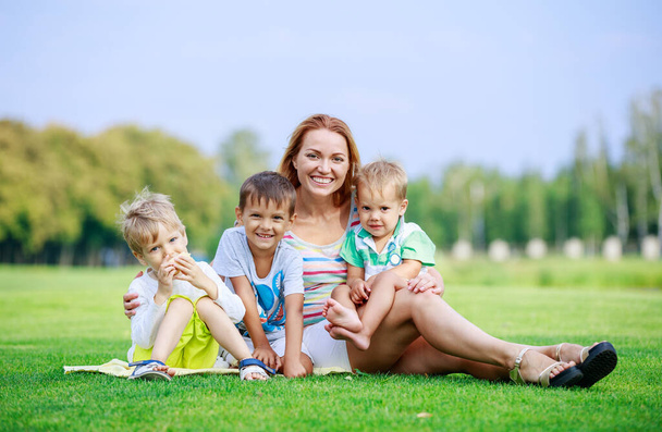 Приваблива молода жінка з маленькими синами сидить на траві в парку. Щаслива сім'я насолоджується яскравим літнім днем. батьківські та дитячі відносини
. - Фото, зображення