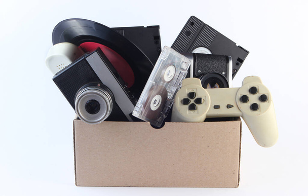 Χάρτινο κουτί με βιντεοκασέτες, ρετρό κάμερα, βινύλιο, κασέτα ήχου, gamepad σε λευκό φόντο. Αντίκες, σκουπίδια - Φωτογραφία, εικόνα