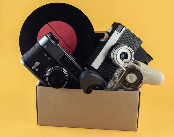 Caixa de papelão com câmera de filme retro, gamepad, registro de vinil, tubo de telefone, cassete de áudio no fundo amarelo. Antiguidades, lixo - Foto, Imagem