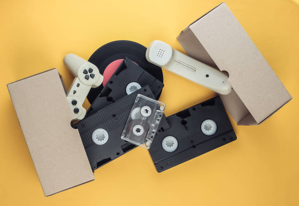 Kartonnen doos met videocassette, retro filmcamera, gamepad, vinylplaat, telefoonbuis, audiocassette op gele achtergrond. Antiek, afval. Bovenaanzicht - Foto, afbeelding