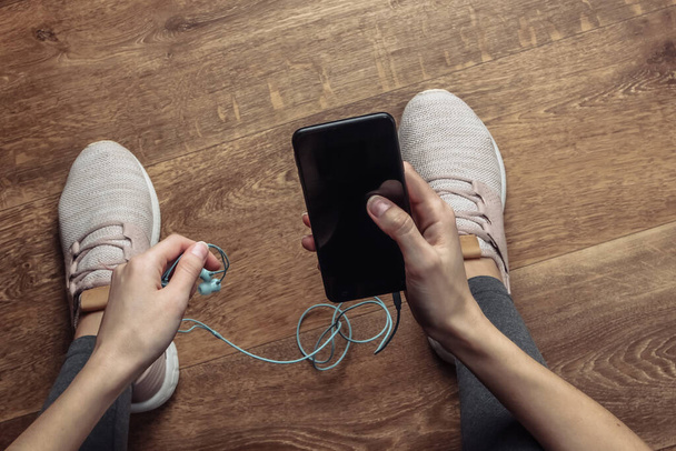 Woman in sportswear sitting on the floor in the room chooses music on smartphone with earphoness for future workout or jogging. Top view - Фото, зображення