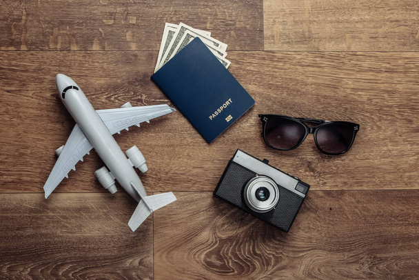 Düz kompozisyonda seyahat et. Uçak heykelciği, kamera, dolar banknotlu pasaport, ahşap zeminde güneş gözlüğü. Dinlenmek, tatil. Üst görünüm - Fotoğraf, Görsel