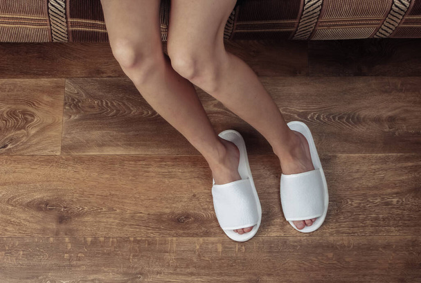 Κάτοψη των γυναικείων γυμνών ποδιών με λευκές παντόφλες σε ξύλινο πάτωμα. Εστία, αφύπνιση - Φωτογραφία, εικόνα