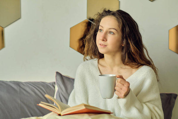 Η γυναίκα κάθεται στο κρεβάτι και πίνει καφέ. απολαμβάνοντας νωρίς το πρωί με ένα βιβλίο στο χέρι. ζεστό πρωινό του χειμώνα. - Φωτογραφία, εικόνα
