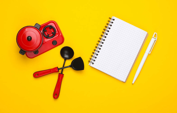 Toy plastic stove with pan, kitchen tool and recipe notebook on yellow background. Chef. Top view, minimalism. Flat lay - Fotoğraf, Görsel