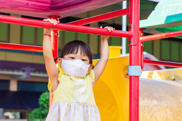 Portraitbild eines 2 bis 3 Jahre alten Babys. Glückliches asiatisches Kindermädchen lächelnd und mit Stoffmaske. Sie zeigt mit dem Finger auf den Spielplatz. Soziale Distanz. Lern- und Aktiv-Konzept für Kinder. - Foto, Bild