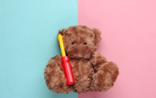 Плюшевий ведмідь тримає іграшкову викрутку на синьо-рожевому пастельному тлі. Концепція ремонту. Вид зверху
 - Фото, зображення