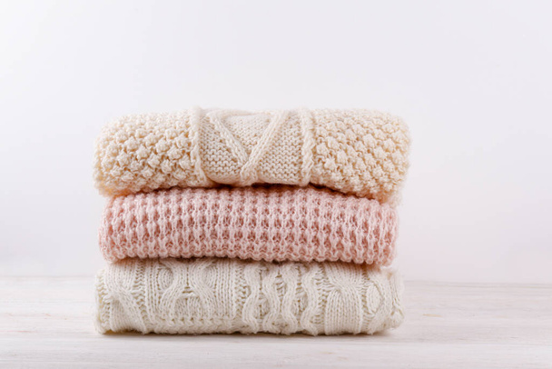 Bunch of knitted warm pastel color sweaters with different knitting patterns folded in stack on white wooden table, white wall background. Fall winter season knitwear. Close up, copy space for text - Photo, image