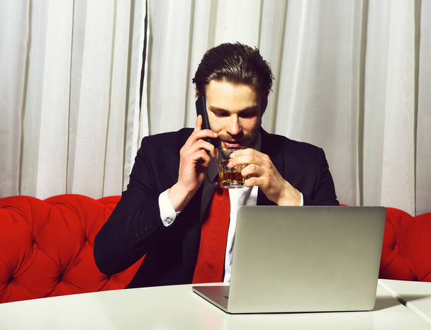 bearded man, businessman holding glass of whiskey works at laptop - Фото, изображение