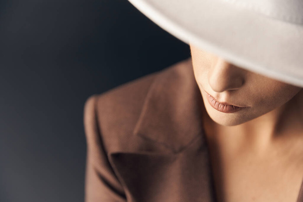 portrait photo on a dark background of a beautiful stylish girl in a brown suit and white hat, where her eyes are not visible - Fotografie, Obrázek