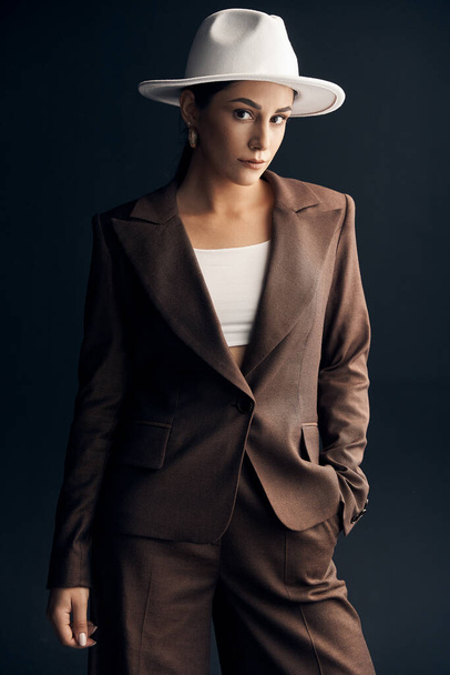 portrait photo on a dark background of a beautiful stylish girl in a brown suit, white top and white hat, she mysteriously looks at the camera with his brown eyes - Фото, зображення