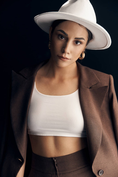 portrait photo of a beautiful stylish girl in a brown suit, white top and white hat, she mysteriously looks into the camera with her brown eyes - Fotografie, Obrázek