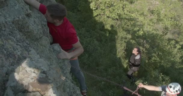 Νεαρός άνδρας σκαρφαλώνει στα βράχια μια ηλιόλουστη μέρα υποστηριζόμενος από την ομάδα - Πλάνα, βίντεο