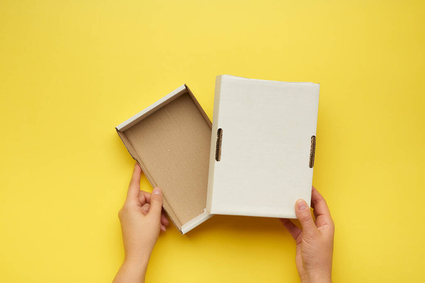 dos manos femeninas sostienen una caja de papel en blanco abierta sobre un fondo amarillo, vista superior - Foto, imagen