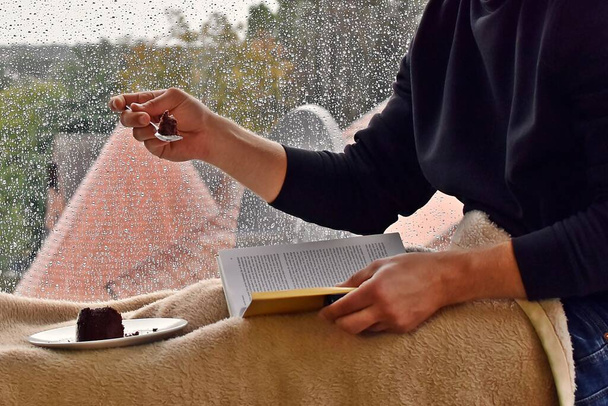 man sitting on a windowsill with delicious chocolate cake, in the background raindrops on the windowpane, man reading a book and enjoying the silence and the rain in autumn - Photo, Image
