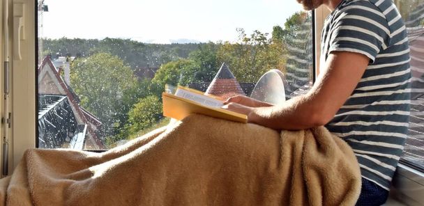 hombre sentado en un alféizar de la ventana, en el fondo gotas de lluvia en el cristal de la ventana, hombre leyendo un libro y disfrutando del silencio y la lluvia en otoño - Foto, imagen