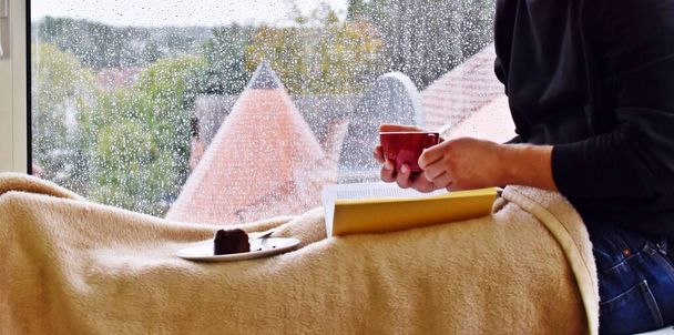 hombre sentado en un alféizar de ventana con taza de bebida caliente, en el fondo gotas de lluvia en el cristal de la ventana, hombre leyendo un libro y disfrutando del silencio y la lluvia en otoño - Foto, Imagen