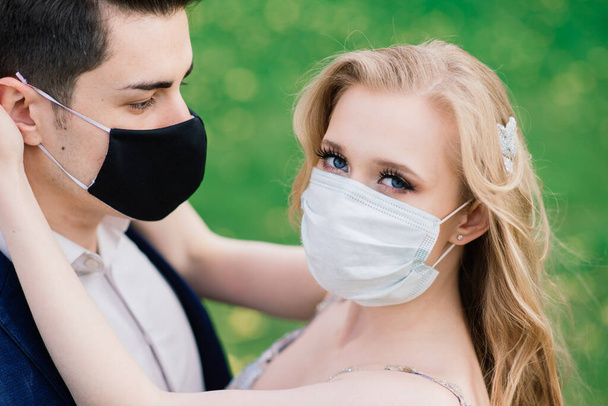 Joven pareja amorosa caminando en máscaras médicas en el parque durante la cuarentena en el día de su boda. - Foto, Imagen