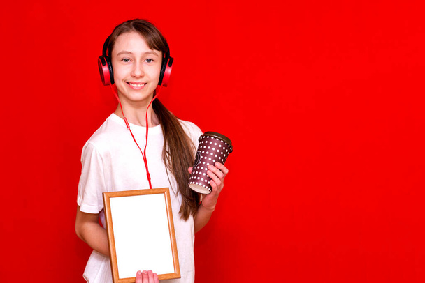 Niedliche kaukasische Brünette mit langen geflochtenen Haaren Schulmädchen in einem weißen T-Shirt mit Cocktail auf einem leuchtend roten Hintergrund. Musik hören, hält leeren Rahmen für ein Banner. - Foto, Bild