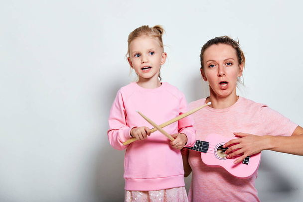 Yong mother and her pretty daughter playing on musical instruments, neutral gray background. Spending funny time together holding ukulele and wood drumsticks. - Photo, Image