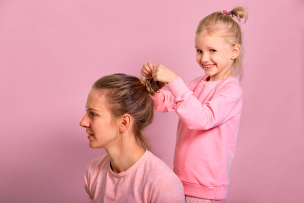 4-5 year old little daughter does the hair of a her mother. Love family spending time together in pink clothes - Fotografie, Obrázek