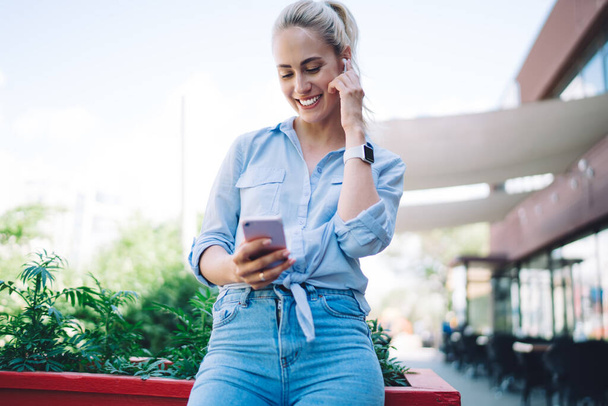 Low angle of happy female in casual clothing smiling and listening to music in wireless earphones while standing on street and messaging on smartphone - Foto, Bild