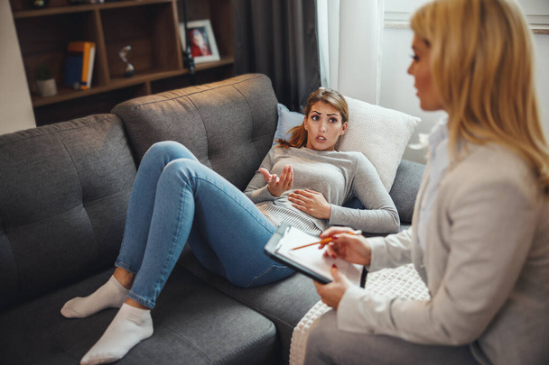Fotografía de una joven preocupada discutiendo con su psicoterapeuta mientras está sentada en un sofá dentro de una sala de estar. - Foto, imagen