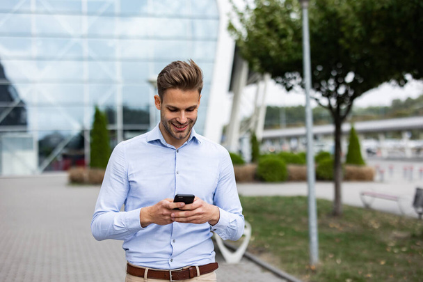Handsome office worker holding smartphone and smiling. Happy young man using mobile phone apps, texting message, browsing internet, looking at smartphone. Coffee break near the business center - Foto, Bild