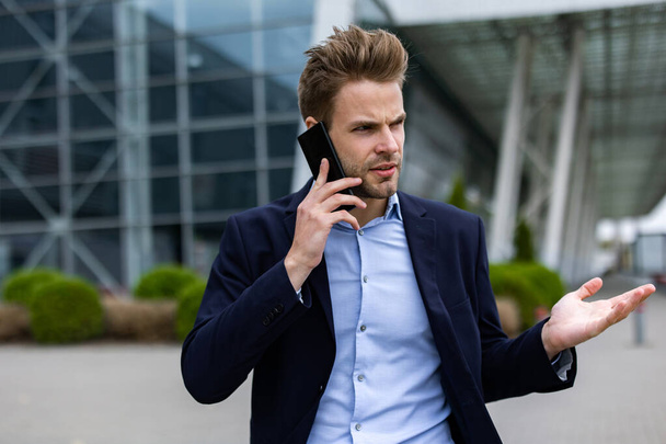 Portrait of handsome smiling man in casual wear holding smartphone. Successful manager makes a call. Young businessman talking on the phone, standing on city street near business center - Foto, immagini