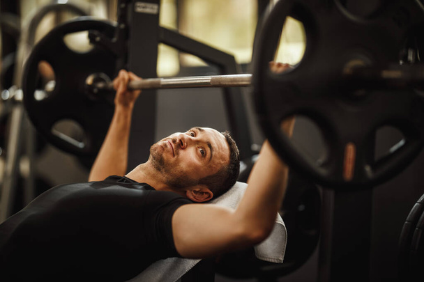 Aufnahme eines muskulösen Kerls in Sportbekleidung beim harten Training in der Sporthalle. Er pumpt mit schwerem Gewicht die Brustmuskulatur auf. - Foto, Bild