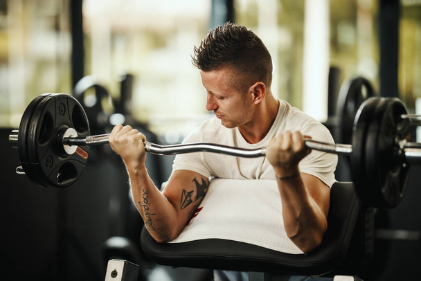 Una foto de un tipo musculoso en ropa deportiva haciendo ejercicio en el duro entrenamiento del gimnasio. Él está bombeando músculo bíceps con peso pesado. - Foto, Imagen