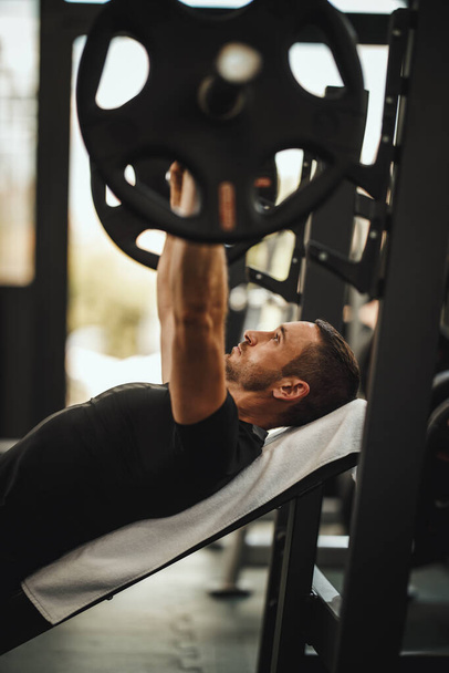 Prise de vue d'un gars musclé en tenue de sport s'entraînant à l'entraînement dur dans la salle de gym. Il pompe la musculature thoracique avec un poids lourd. - Photo, image