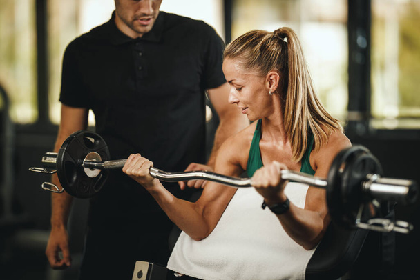 Aufnahme einer muskulösen jungen Frau in Sportbekleidung, die mit Personal Trainer im Fitnessstudio trainiert. Sie pumpt ihre Bizeps-Muskulatur mit schwerem Gewicht auf. - Foto, Bild