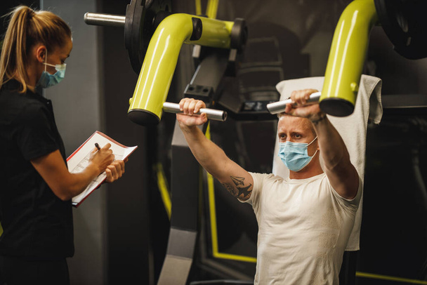 Foto de una joven musculosa con máscara protectora que trabaja con un entrenador personal en la máquina de gimnasia durante la pandemia de Covid-19. Ella está bombeando su músculo torácico con peso pesado. - Foto, Imagen