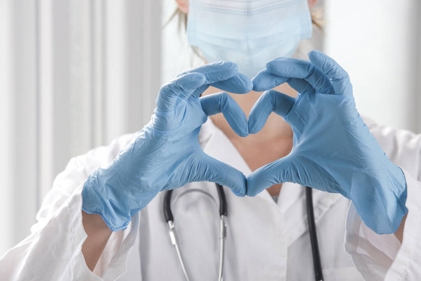 Zbliżenie wizerunku pracownika medycznego noszącego ochronną maskę chirurgiczną i gumowe rękawiczki sprawia, że znak serca dłońmi. - Zdjęcie, obraz