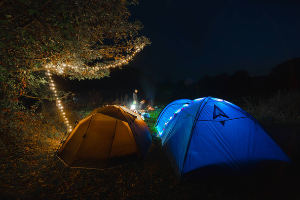 Touristisches Zeltlager. Girlanden, die leuchten. Eine Nacht in einer Wildnis. Menschen im Urlaub. Aktives Lifestylekonzept und Ausrüstung für Freizeit und Wandern. Klappstühle und Tisch. - Foto, Bild