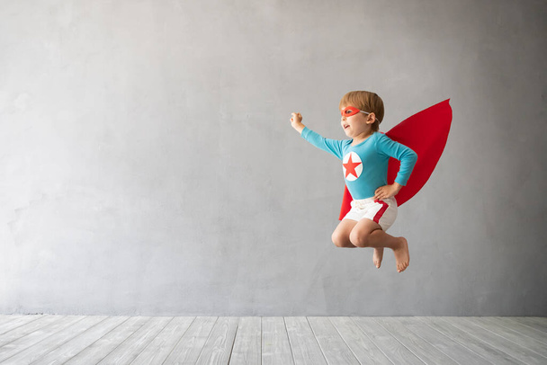 Superhero child jumping against grey concrete background. Super hero kid playing at home. Childhood dream and freedom concept - Photo, image