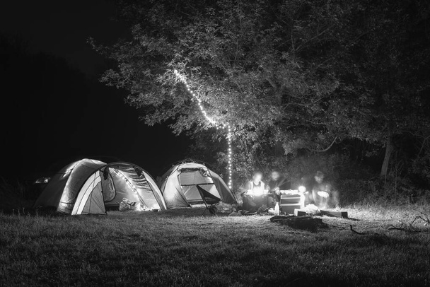 Tourist camping tent garlands, lighting up. A night in a wilderness place. People on vacation. Active lifestyle concept and equipment for recreation hiking. folding chairs and table. black and white - Photo, Image