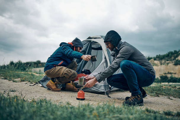 雨の日に二人の男がテントの近くでうずくまってコーヒーを準備した。キャンプ旅行のコンセプト. - 写真・画像