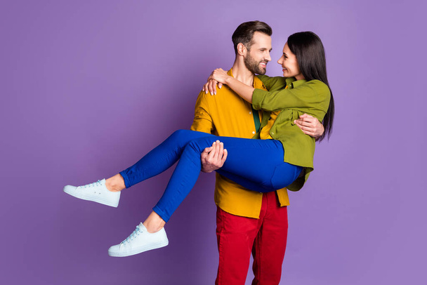 Photo pretty lady handsome guy couple carry wife bride hold arms playful mood wedding day look eyes tempting wear casual bright shirts pants outfit isolated purple color background - Foto, afbeelding