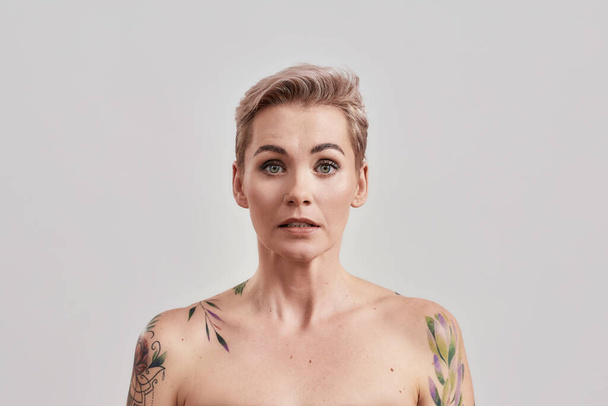 Oops. Portrait of half naked tattooed woman with short hair feeling awkward, silly isolated over light background - Photo, Image