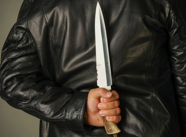 a man in a black leather jacket holds a large knife behind his back - Fotoğraf, Görsel