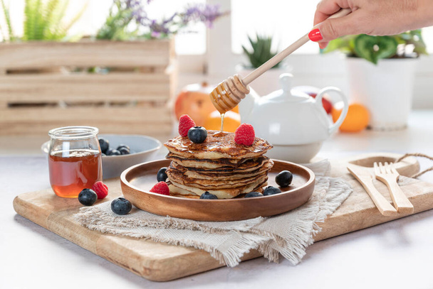 Homemade pancakes with fresh blueberries, raspberries and honey. Female hand pouring honey - Photo, Image